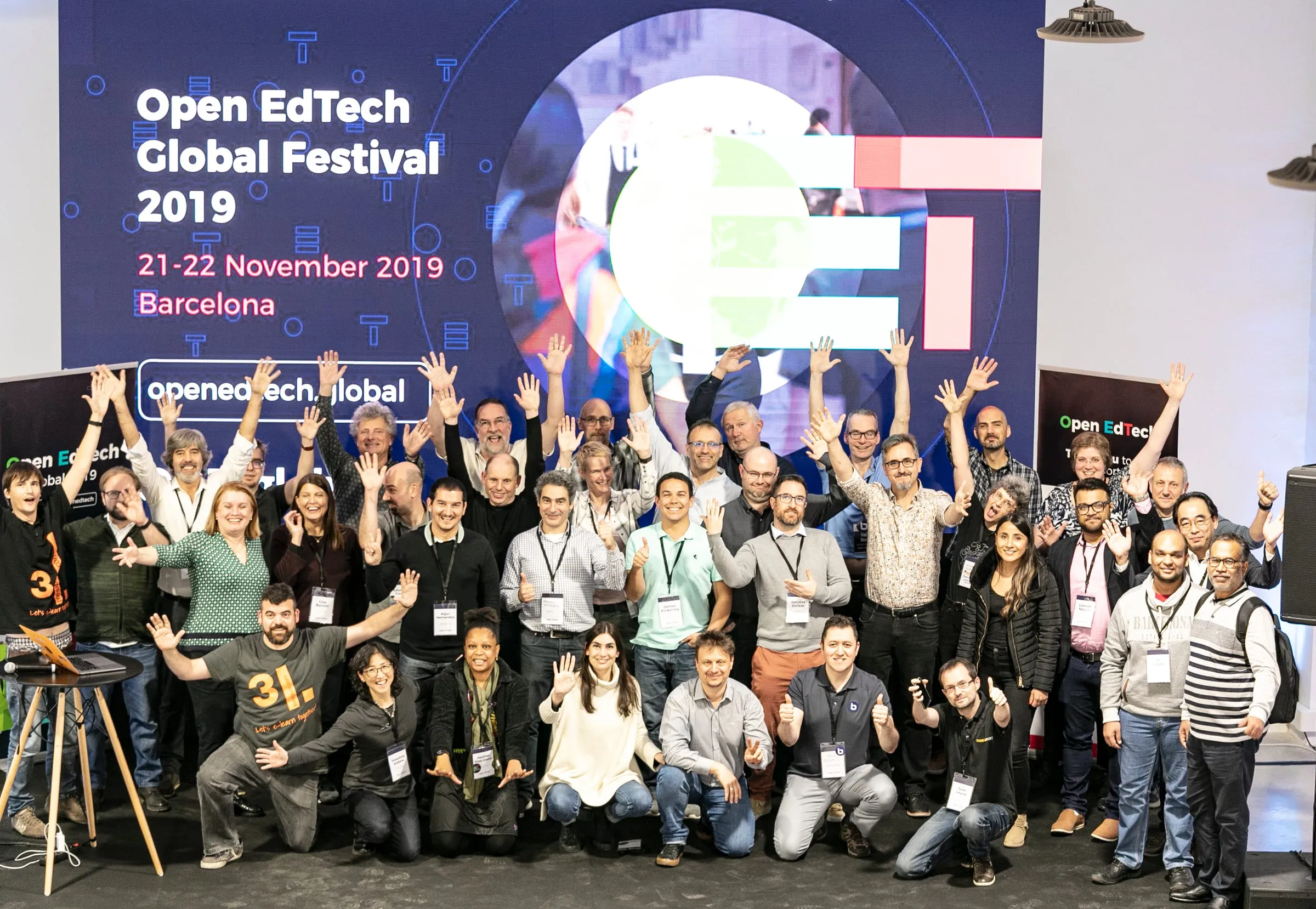 About forty people posing with their arms raised in front of a large screen reading Open EdTech Global Festival 2019, 21 to 22 November 2019, Barcelona.
